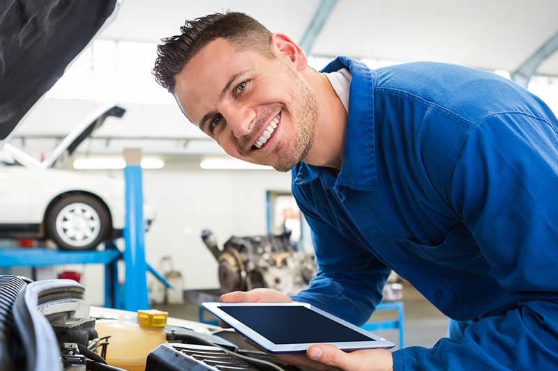 Smiling technician servicing a car engine while holding a tablet.